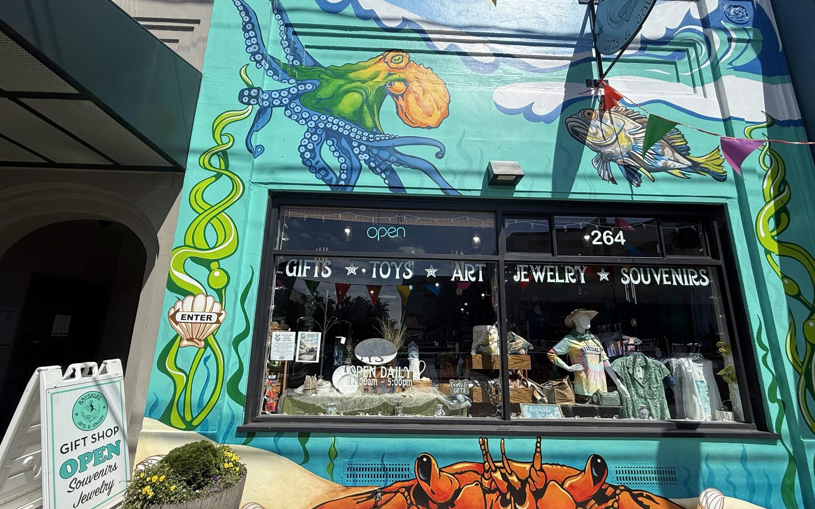Bayshore Gifts and Gallery entrance with a painted octopus and kelp mural, a scallop shell enter sign, and an open gift shop sign